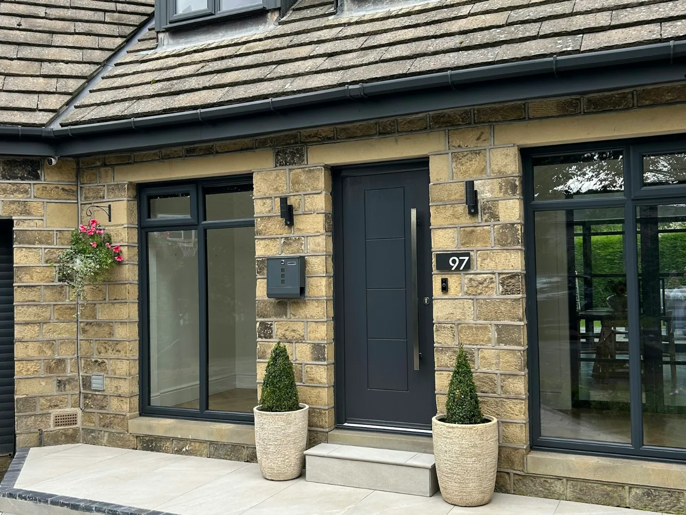 Porte d'entrée gris anthracite à barre de tirage, entre deux grandes fenêtres fixes, sur une maison en pierre